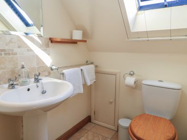 A bathroom with a white sink and chrome taps a toilet with a wooden seat a small shelf with toilet paper and towels hanging on a rail under a skylight window at Granary Cottage in Blackwell near Ilmington
