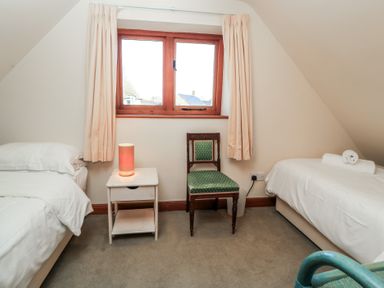A bedroom with two single beds a nightstand with a lamp a green cushioned wooden chair and a window with curtains at Granary Cottage in Blackwell near Ilmington