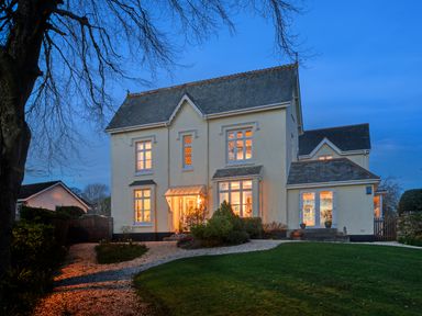 A large two-story house with lit windows and a gravel driveway surrounded by trees and lawn at Victoria Lodge in Kingsbridge