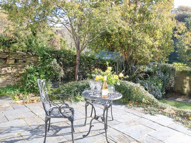 A patio area with a metal table holding a bottle and glasses and a metal chair surrounded by trees and plants at Victoria Lodge in Kingsbridge