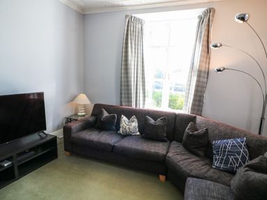 A living room with a dark sectional sofa and cushions near a window with curtains and a television on a stand at Victoria Lodge in Kingsbridge