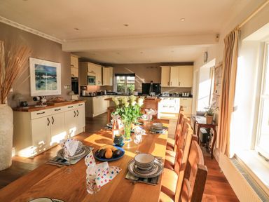 A dining area with a wooden table set with dishes and napkins and a kitchen with cream cabinets in the background at Victoria Lodge in Kingsbridge