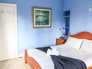 A bedroom with a wooden bed and white linens a nightstand with a lamp and a painting on the blue wall at Victoria Lodge in Kingsbridge