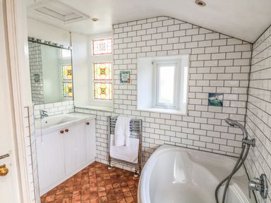 A bathroom with white tiled walls a corner bathtub a sink cabinet a towel radiator and stained glass windows at Victoria Lodge in Kingsbridge