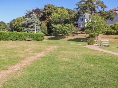 A grassy park area with a wooden bench a pathway and a white house surrounded by trees at Victoria Lodge in Kingsbridge