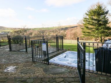 An outdoor space with a fenced hot tub stone patio and grassy area with hills and wind turbines in the background at Nant Moel Isaf Farm in Rhydyfro near Pontardawe