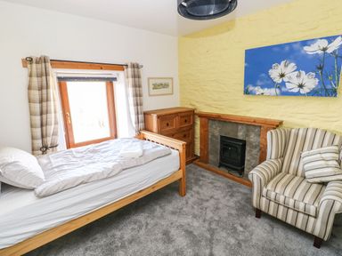 A bedroom with a single bed, wooden chest of drawers, striped armchair, fireplace, and window with plaid curtains at Nant Moel Isaf Farm in Rhydyfro near Pontardawe