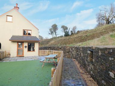 An outdoor area with a small fenced yard artificial grass wooden picnic table stone pathway and a beige house with wooden windows at Nant Moel Isaf Farm in Rhydyfro near Pontardawe