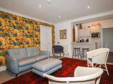 A living room with a grey sofa and ottoman two white wicker chairs and a blue armchair next to a kitchen with two stools with a floral wallpaper on one wall at Melvill Lower Deck in Falmouth