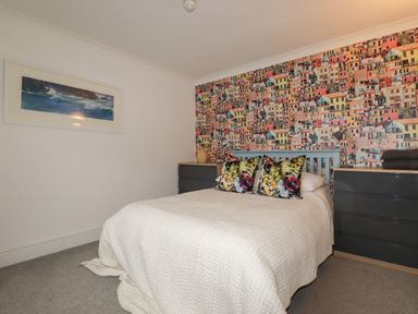 A bedroom with a bed covered with a white quilt and colorful pillows and two dark dressers with lamps on each side and a wall with house pattern wallpaper at Melvill Lower Deck in Falmouth