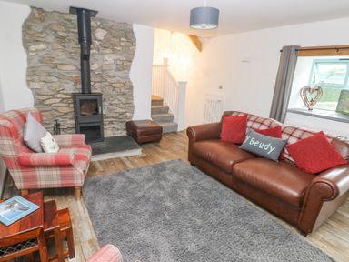 A living room with a stone fireplace wood stove a leather sofa with red cushions a plaid armchair a grey rug and stairs leading up at Y Beudy Crosswell Eglwyswrw near Newport Pembrokeshire