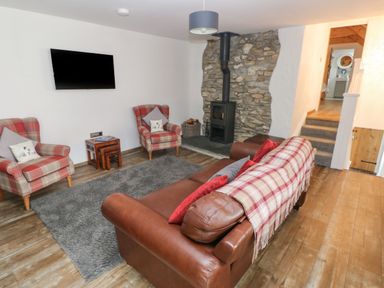 A living room with leather sofa, two plaid armchairs, a wall-mounted TV, a wood stove on a stone wall and a gray rug at Y Beudy in Crosswell, Eglwyswrw near Newport, Pembrokeshire