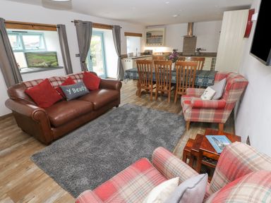 A living area with a brown leather sofa, plaid armchairs, a wooden dining table with chairs, and a kitchen in the background at Y Beudy in Crosswell Eglwyswrw near Newport Pembrokeshire
