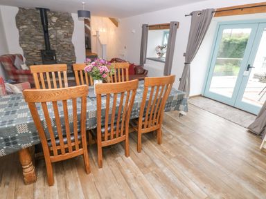 A dining area with wooden chairs and a table with a tablecloth and flowers a seating area with armchairs and a wood burning stove near a stone wall and glass doors leading outside at Y Beudy Crosswell Eglwyswrw near Newport Pembrokeshire