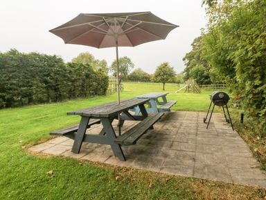 An outdoor patio area with two wooden picnic tables one with an umbrella and a barbecue grill with a grassy field and playground equipment in the background at Y Beudy in Crosswell Eglwyswrw near Newport Pembrokeshire