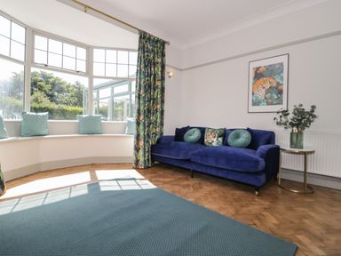A living room with a blue sofa and round cushions a bay window with cushions floral curtains and a side table with plant at Monarfon in Amlwch