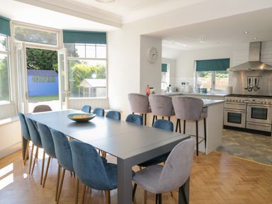 A dining area with a long table and blue chairs next to an open door and a kitchen with bar stools and a stove at Monarfon in Amlwch