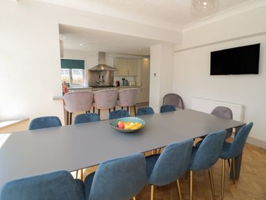 A dining area with a large table surrounded by blue chairs and three bar stools at a kitchen counter with beige cabinets at Monarfon in Amlwch