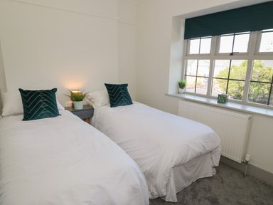 A bedroom with two single beds with white bedding and green pillows a side table with a lamp and plant and a window with plants on the sill at Monarfon in Amlwch