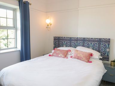 A bedroom with a bed with a patterned headboard and pink pillows blue curtains and a bedside table with a plant at Monarfon in Amlwch