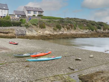 A shoreline with three kayaks and one inflatable boat on a rocky beach and houses on a hill at Monarfon in Amlwch