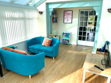 A sunlit room with two blue armchairs with cushions a wooden table with a TV a bookshelf and a glass door at Monarfon in Amlwch