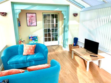 A room with turquoise sofas and orange cushions a wooden coffee table with a television a basket with items vertical blinds and glass doors at Monarfon in Amlwch