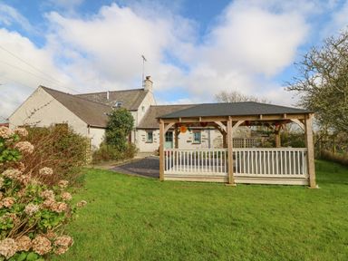 A garden with a wooden gazebo and a stone house in the background at Ty Gwennol in Llanfaethlu