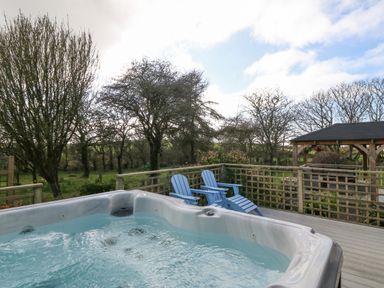 An outdoor deck with a hot tub two blue chairs trees and a wooden gazebo at Ty Gwennol in Llanfaethlu