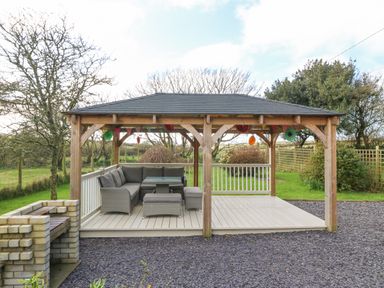A wooden gazebo with outdoor seating and a table on a deck next to a brick barbecue in a garden at Ty Gwennol in Llanfaethlu