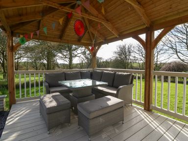 An outdoor covered seating area with a corner sofa, two ottomans, a glass-topped table, and decorative hanging flags at Ty Gwennol in Llanfaethlu