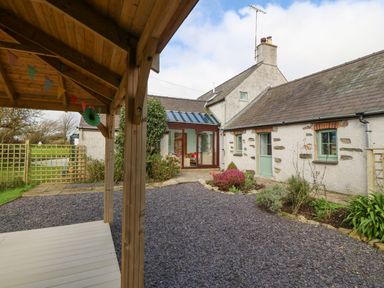 An outdoor area with a gravel path a wooden covered porch and a stone building with green doors and windows at Ty Gwennol in Llanfaethlu