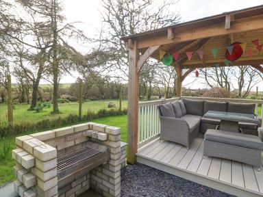 An outdoor seating area with a table under a wooden gazebo and a brick barbecue grill in a garden at Ty Gwennol in Llanfaethlu