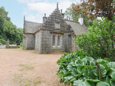 A stone building with pointed roof and lattice windows surrounded by trees and plants at East Lodge Crathes Castle near Banchory