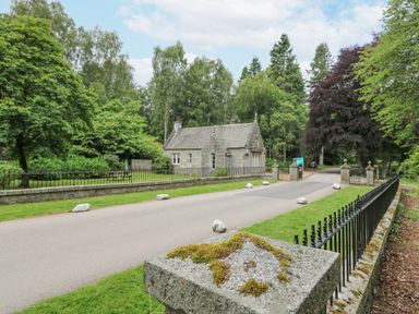 A stone building behind a metal fence along a paved road surrounded by trees at East Lodge - Crathes Castle in Crathes Castle near Banchory