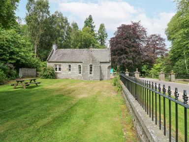 A small stone house with a grassy lawn and a picnic table beside a road with a black metal fence at East Lodge Crathes Castle near Banchory