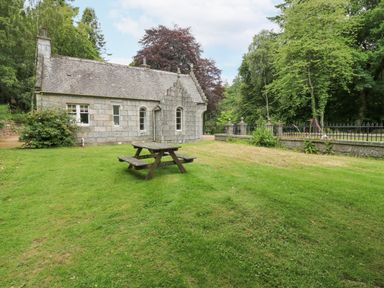 A stone house with a sloped roof and arched windows on a grassy lawn with a wooden picnic table at East Lodge - Crathes Castle in Crathes Castle near Banchory