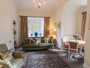 A living room with a sofa and armchair with cushions and a dining table with flowers at East Lodge - Crathes Castle in Crathes Castle near Banchory