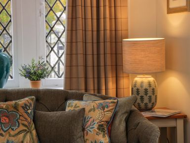 A corner of a living room with a patterned sofa cushions a table lamp on a side table and a potted plant on a window sill at East Lodge Crathes Castle near Banchory
