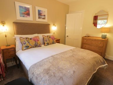 A bedroom with a double bed with floral pillows two bedside tables a chest of drawers with a lamp and mirror above at East Lodge - Crathes Castle in Crathes Castle near Banchory