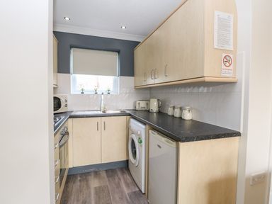 A kitchen with light wood cabinets a black countertop white appliances and a window with a blind at 91 Waterside Park in Lowestoft