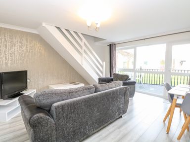 A living room with grey sofas a TV white tables a dining table and chairs and a staircase at 91 Waterside Park in Lowestoft
