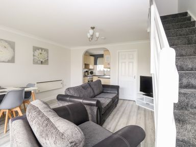 A living room with grey sofas a white dining table and chairs a TV on a stand and a staircase at 91 Waterside Park in Lowestoft