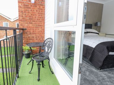 A small balcony with a black metal table and chair set a potted plant on artificial grass and a bedroom with a bed and lamp visible through an open door at 91 Waterside Park in Lowestoft