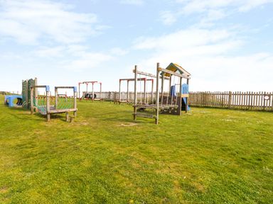 An outdoor playground with climbing frames swings and a slide on grass surrounded by a wooden fence at 91 Waterside Park in Lowestoft