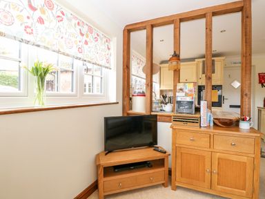 A corner of a room with a television on a wooden stand and a wooden cabinet with guest information and kitchen visible through wooden framed window at Hunters Lodge Great Moulton near Long Stratton