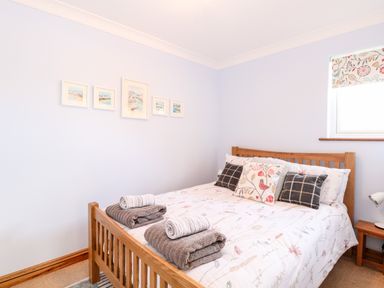 A bedroom with a wooden bed frame, patterned bedspread, pillows, folded towels on the bed, framed pictures on the wall and a small window with floral blind at Hunters Lodge in Great Moulton near Long Stratton