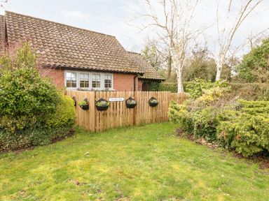 A garden with grass shrubs and a wooden fence with flower pots in front of a brick house at Hunters Lodge Great Moulton near Long Stratton