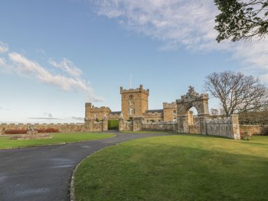 A castle with a clock tower a stone archway and a curved paved driveway surrounded by green grass at Ardlochan Lodge - Culzean Castle in Maybole
