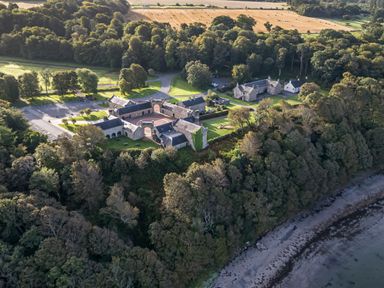 An aerial view of a historic building complex surrounded by trees and fields near a shoreline at Ardlochan Lodge - Culzean Castle in Maybole